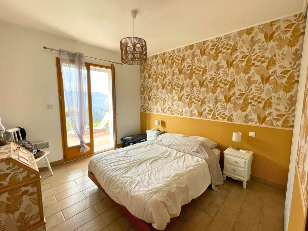 Chambre rénovée avec papier peint végétal, sous-bassement ocre et mobilier assorti, ambiance chaleureuse et naturelle.