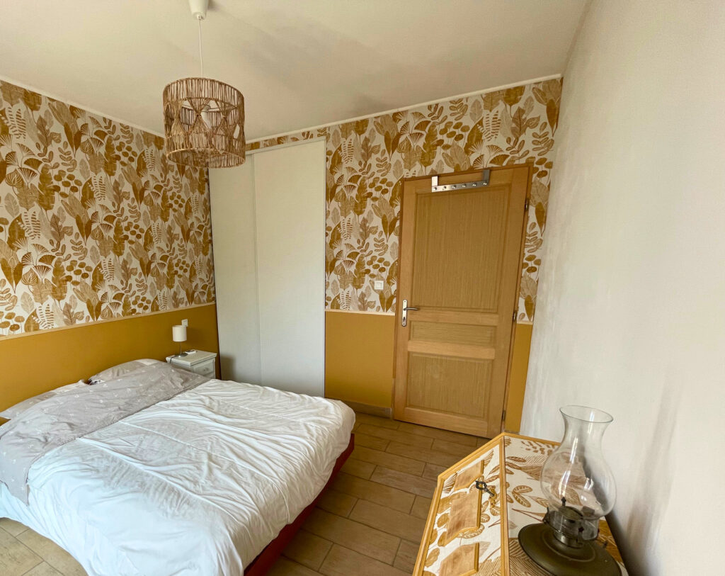 Chambre rénovée avec papier peint végétal, sous-bassement ocre et mobilier assorti, ambiance chaleureuse et naturelle.