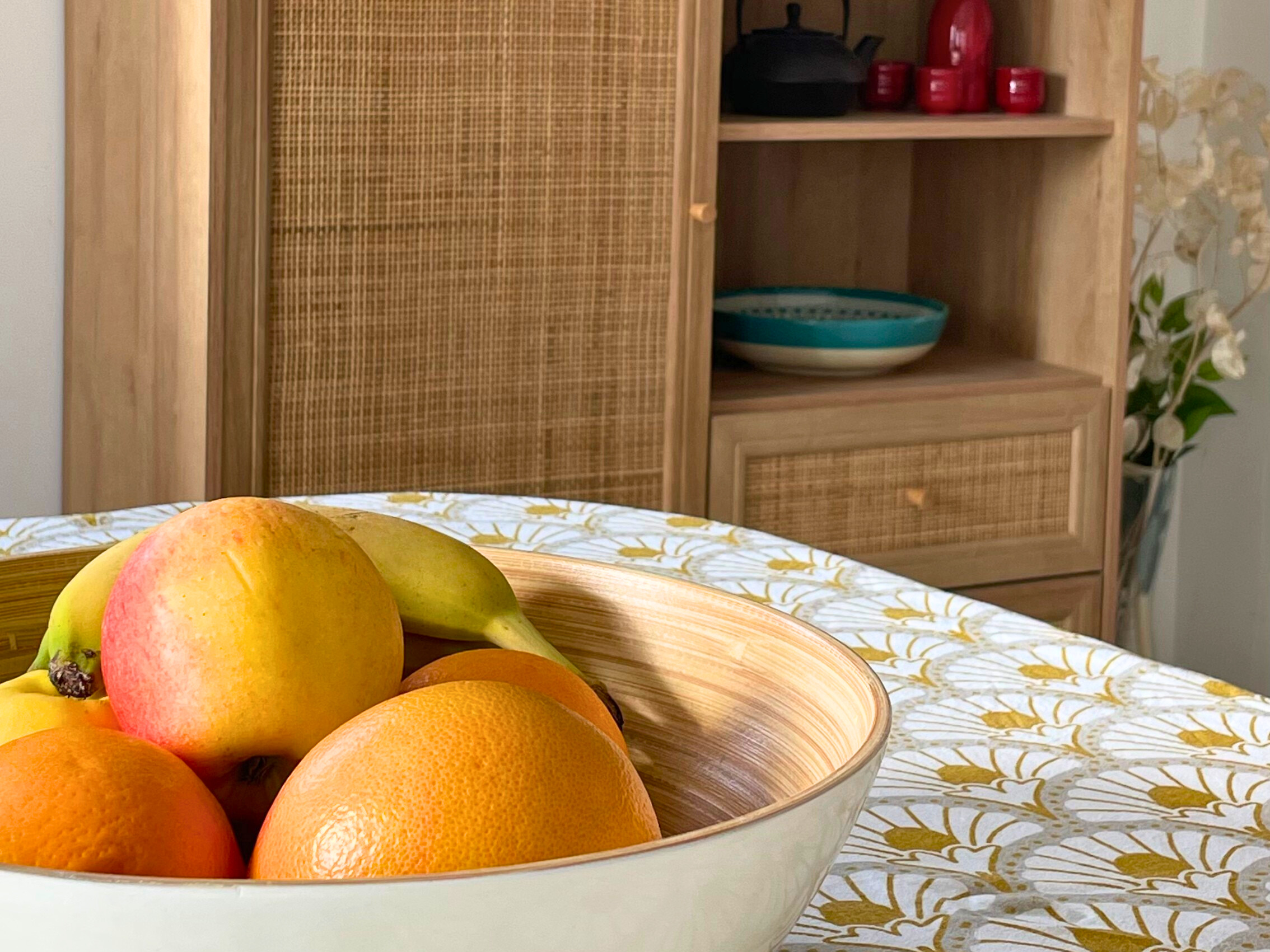 Détails de matière, un coin de table avec nappe et coupe de fruits au premier plan, meuble bois et porte en rotin à l'arrière plan