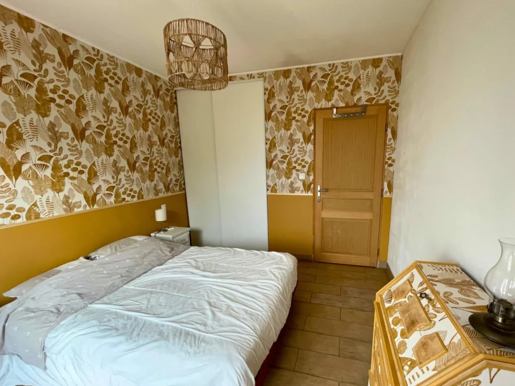 Chambre rénovée avec papier peint végétal, sous-bassement ocre et mobilier assorti, ambiance chaleureuse et naturelle.